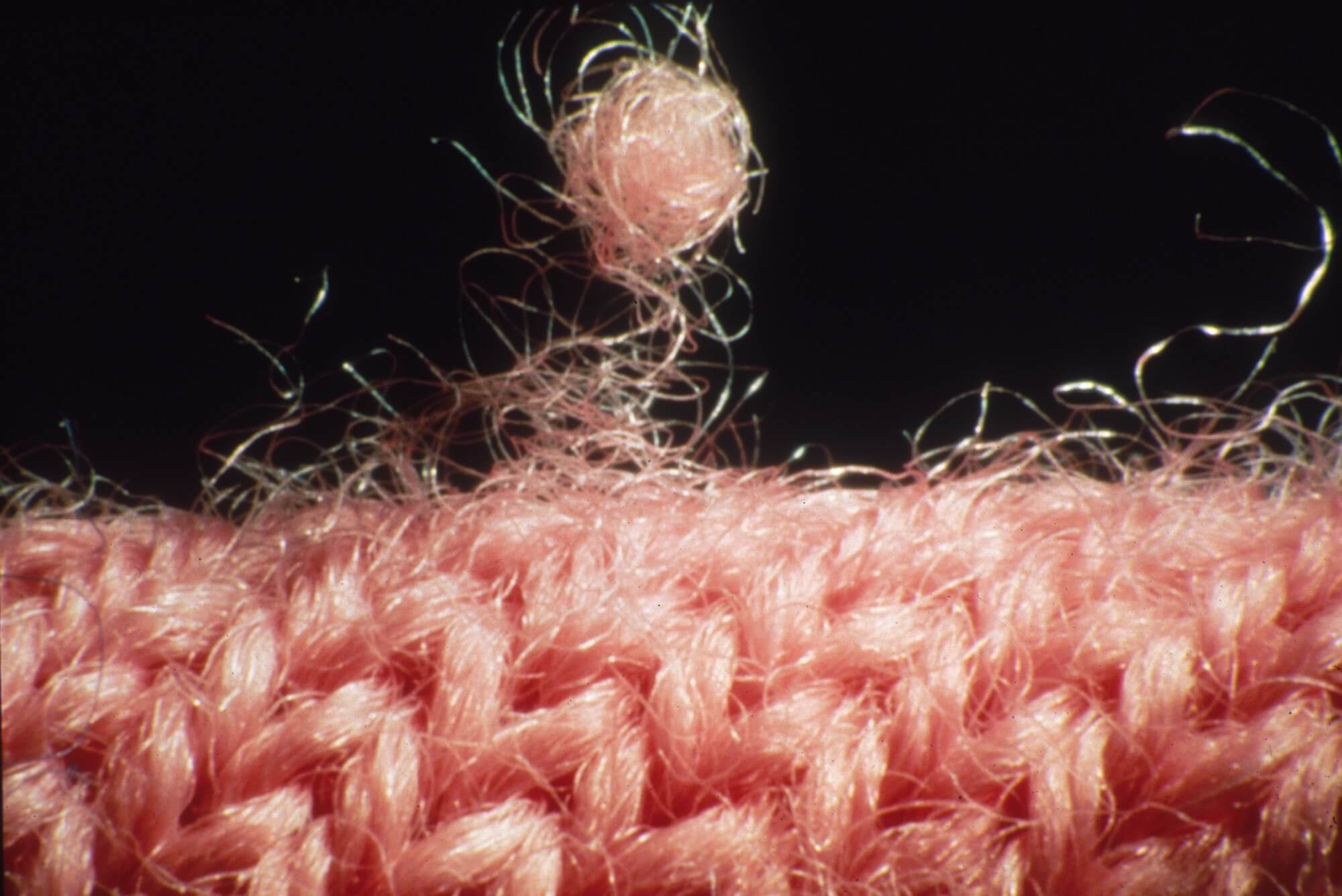 A macro close-up shot of some kind of pink wool. There are many loose threads, and one large piece of lint seemingly about to come off the fabric.